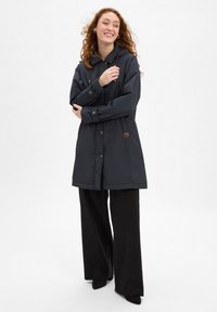 Navy outerwear jacket with a hood, featuring gold-tone hardware, a logo patch, and a long, straight fit, paired with black wide-leg pants.
