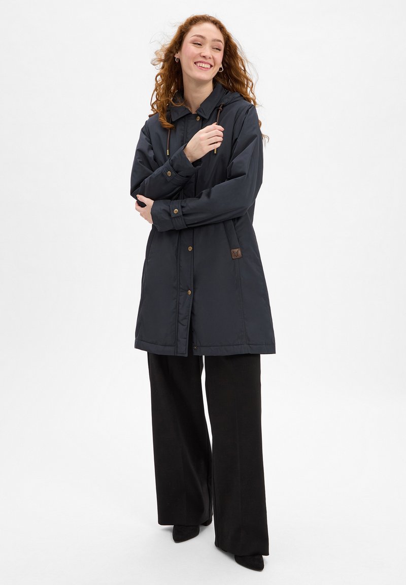 Navy outerwear jacket with a hood, featuring gold-tone hardware, a logo patch, and a long, straight fit, paired with black wide-leg pants.