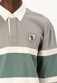 Grå og grøn stribet polo skjorte med en hvid krave og et våbenskjold, der har en løve. Stoffet ser blødt ud med en struktureret overflade.
