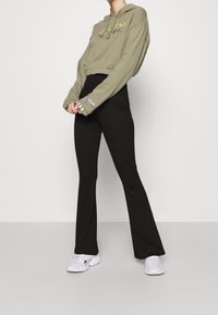 Person in cropped olive Adidas hoodie, black high-waisted flared pants, and white sneakers standing against plain light background.