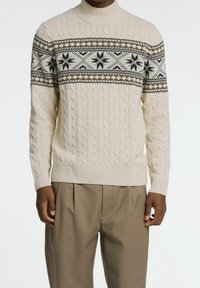 Stickad tröja i krämfärg med kabelstruktur, med en mörk mönstrad ok med snöflingeformer. Hög hals och ribbade ärmslut.