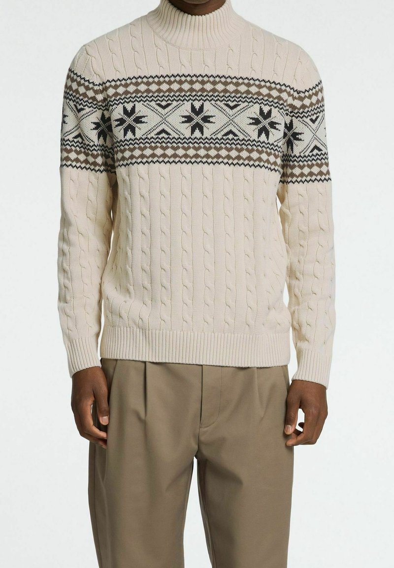 Stickad tröja i krämfärg med kabelstruktur, med en mörk mönstrad ok med snöflingeformer. Hög hals och ribbade ärmslut.