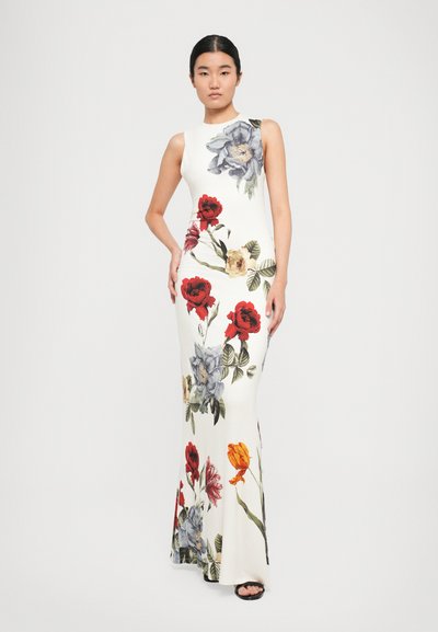 Woman stands wearing a sleeveless white floor-length dress adorned with large colorful floral prints and black open-toe sandals.