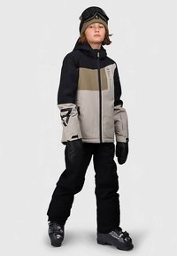 Junger Mensch, der eine schwarze, beige und olivfarbene Skijacke, schwarze Schneehosen, Handschuhe, eine Mütze, eine Skibrille und Skischuhe trägt und vor einem einfarbigen Hintergrund steht.