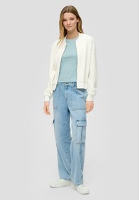 Witte textured bomberjack, lichtblauw fitted top, blauwe cargo jeans met zakken en witte sneakers. Model staat tegen een neutrale achtergrond.
