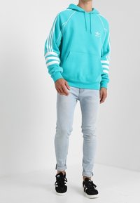 Turkos hoodie med vit tre-randig design på ärmarna, känguruficka och dragsko i huvan, kombinerad med ljusblå jeans och svarta sneakers.