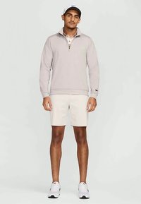 Lys grå zip-up genser med krage og lange ermer, kombinert med hvite shorts. Modellen har på seg hvite joggesko og en matchende caps.