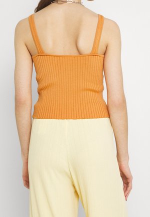Femme portant un haut orange sans manches côtelé et un pantalon plissé jaune clair, vue de dos sur un fond uni.