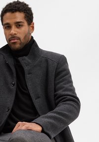 Dunkelgrau Wollmantel mit hohem Kragen, ausgestattet mit Knopfverschlüssen und einer strukturierten Oberfläche; getragen über einem schwarzen Rollkragenpullover.