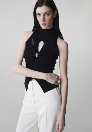 Donna con capelli scuri che indossa un top nero senza maniche con dettaglio centrale ad anello oro e pantaloni bianchi a vita alta, su uno sfondo neutro.