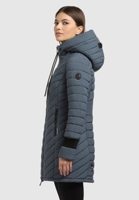 Sivá prešívaná dlhá bunda s kapucňou, čierne akcenty, vrecká na zips a logo nášivka na rukáve. Disponuje štýlovým dizajnom.