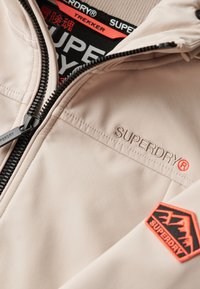 Lichtbeige jack met zwarte ritssluiting, geborduurd "SUPERDRY" logo en oranje bergpatch. Soepele stof, sportief ontwerp met capuchon.