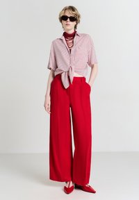 Rote Schlaghosen kombiniert mit einer gestreiften, gebundenen Bluse. Die Accessoires umfassen eine dunkle Sonnenbrille und eine mehrlagige rote Halskette. Sanfte Texturen.