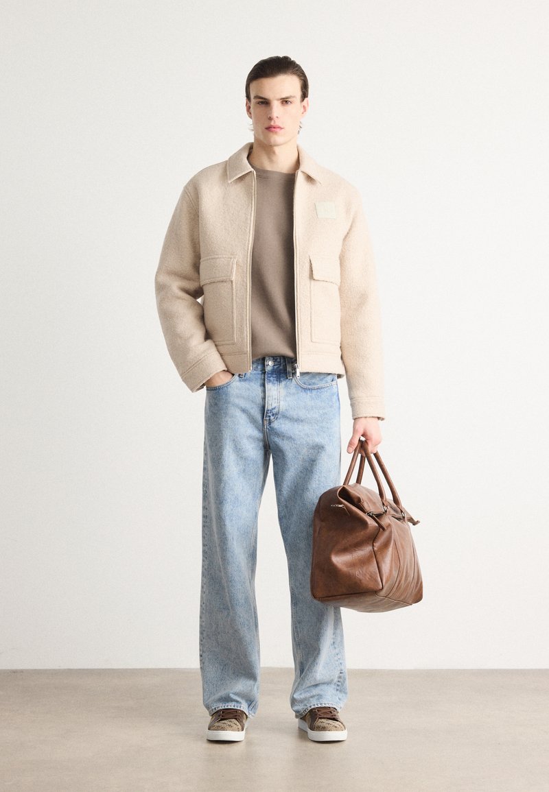 Chaqueta de mezcla de lana color beige con dos bolsillos frontales, camisa topo debajo, jeans de pierna ancha azul claro, bolso de cuero marrón y zapatillas grises.