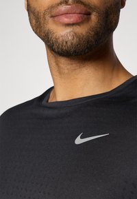 Black athletic t-shirt with a round neckline, featuring a silver Nike logo on the chest and subtle textured patterns across the fabric.