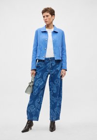 Μπλε υφασμάτινο μπουφάν με γιακά, δύο μπροστινές τσέπες, συνδυασμένο με ψηλόμεσα τζιν παντελόνια με σχέδιο πεϊσλί. Μαύρα μποτάκια μέχρι τον αστράγαλο και γκρι τσάντα.