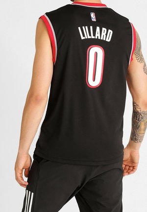 Mężczyzna ubrany w czarną koszulkę do koszykówki z czerwonym wykończeniem, z napisem „Lillard” i numerem „0” na plecach, z tatuażem na prawej ręce i czarnymi sportowymi spodniami.