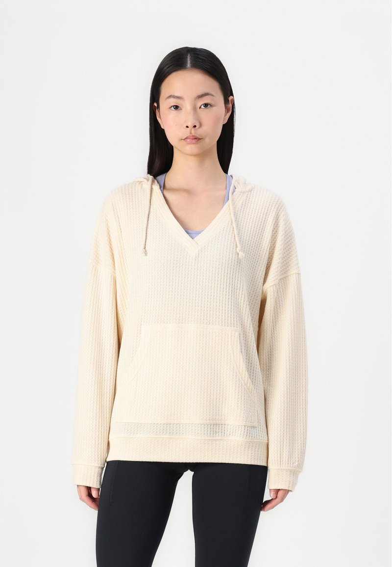 Cremefarbener Kapuzenpullover mit V-Ausschnitt, strukturiertem Stoff, Fronttasche und langen Ärmeln, kombiniert mit schwarzen Leggings.