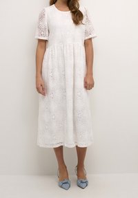 Valkoinen pitsinen midi-mekko, jossa on lyhyet puffihihat, kukkakuviot ja liehakka helma. Yhdistetty vaaleansiniin nauhoilla koristeltuihin korkokenkiin.