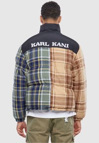 Pufjakke med et flerfarvet ternet design i grøn, blå, brun og beige, med en sort krave og "KARL KANI" logo på bagsiden.