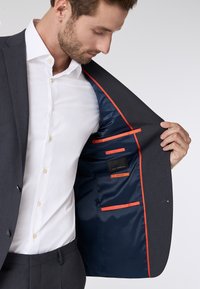 Donkergrijs colbert met een gladde textuur, voorzien van een marineblauwe voering en oranje accenten. Binnenzakken tonen labels en levendige details.