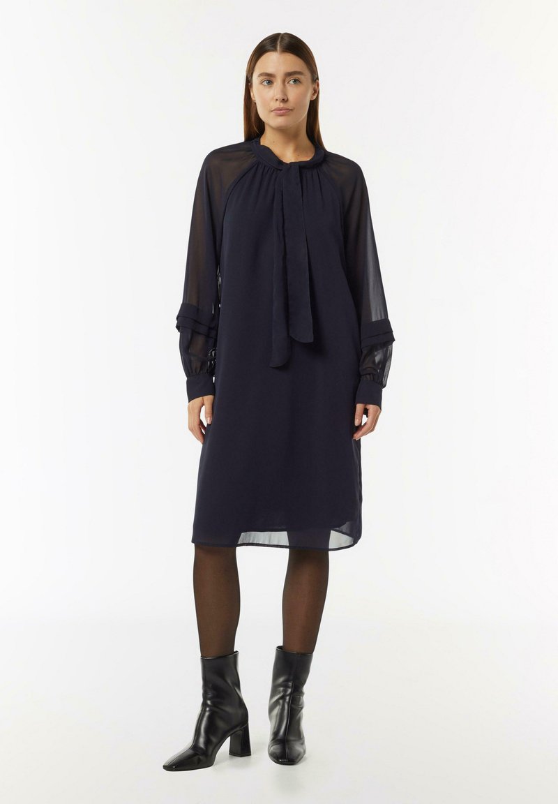 Navyblaues Kleid mit durchsichtigen langen Ärmeln, einem gerafften Ausschnitt und einem Bindeband vorne; kombiniert mit schwarzen Ankle Boots.