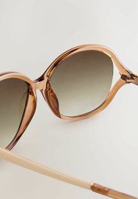 Des lunettes de soleil avec une monture brillante de couleur brun clair, des verres à dégradé sombre et une forme incurvée. La branche est de couleur beige clair.