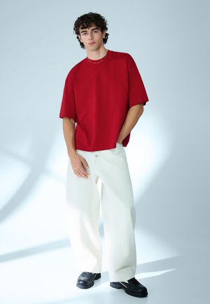 Giovane uomo in piedi con una mano in tasca, indossa una maglietta rossa oversize, pantaloni bianchi larghi, scarpe nere e una sottile collana d'oro su sfondo bianco.