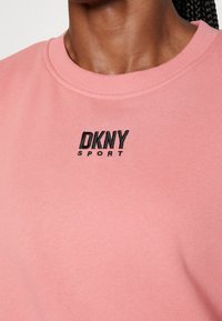 Sweatshirt rose avec un design à col rond. Broderie noire "DKNY SPORT" affichée sur la poitrine gauche. Texture de tissu douce.