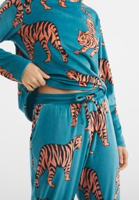 Conjunto de loungewear de felpa azul con un patrón de tigres naranjas por todo el tejido. La parte superior tiene un corte relajado y los pantalones cuentan con una cintura ajustable con cordón.