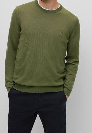 Mężczyzna w oliwkowozielonym swetrze z okrągłym dekoltem z małym logo "BOSS" na piersi i czarnych spodniach, z rękami w kieszeniach, na jednolitym tle.