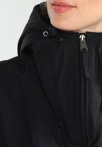 Czarna kurtka z zamkiem z przodu, regulowanym kapturem ze sznurkiem i gładkim, lekkim materiałem o matowym wykończeniu.