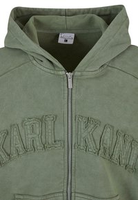 Zeleni zip-up dukserica s teksturiranim logotipom "KARL KANI" na prsima. Ima kapuljaču, prednji zip i mekanu tkaninu.
