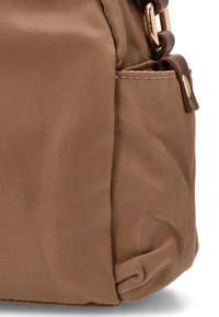 Bolso de tela marrón con una textura suave, que cuenta con un bolsillo lateral, herrajes en tono dorado y bordes suaves y redondeados. Diseño compacto.