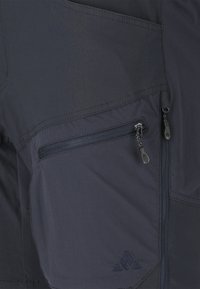 Nærbillede af mørke stofbukser med to lynlåslommer, synlige syninger og et lille broderet logo på den nederste lomme.