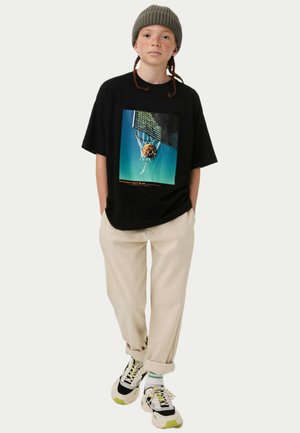 Zwarte T-shirt met een grafische print van een basketball in een net. Draagt het met beige broeken en zwarte, witte en groene sneakers.