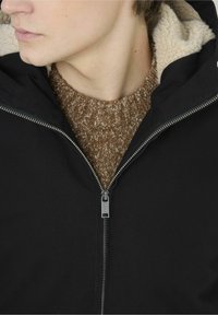 Veste noire zippée avec une douce doublure en fausse fourrure crème, portée sur un pull en tricot brun texturé. Détail de la fermeture éclair et du col visible.