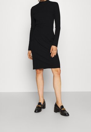 Femme portant une robe noire cintrée à manches longues et longueur genou, ainsi que des mocassins noirs à talons avec un détail chaîne dorée, debout contre un mur uni.