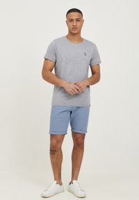 Tricou gri cu mânecă scurtă și guler rotund, având un mic logo brodat; asociat cu pantaloni scurți bleu cu mansete rulate și adidași albi.