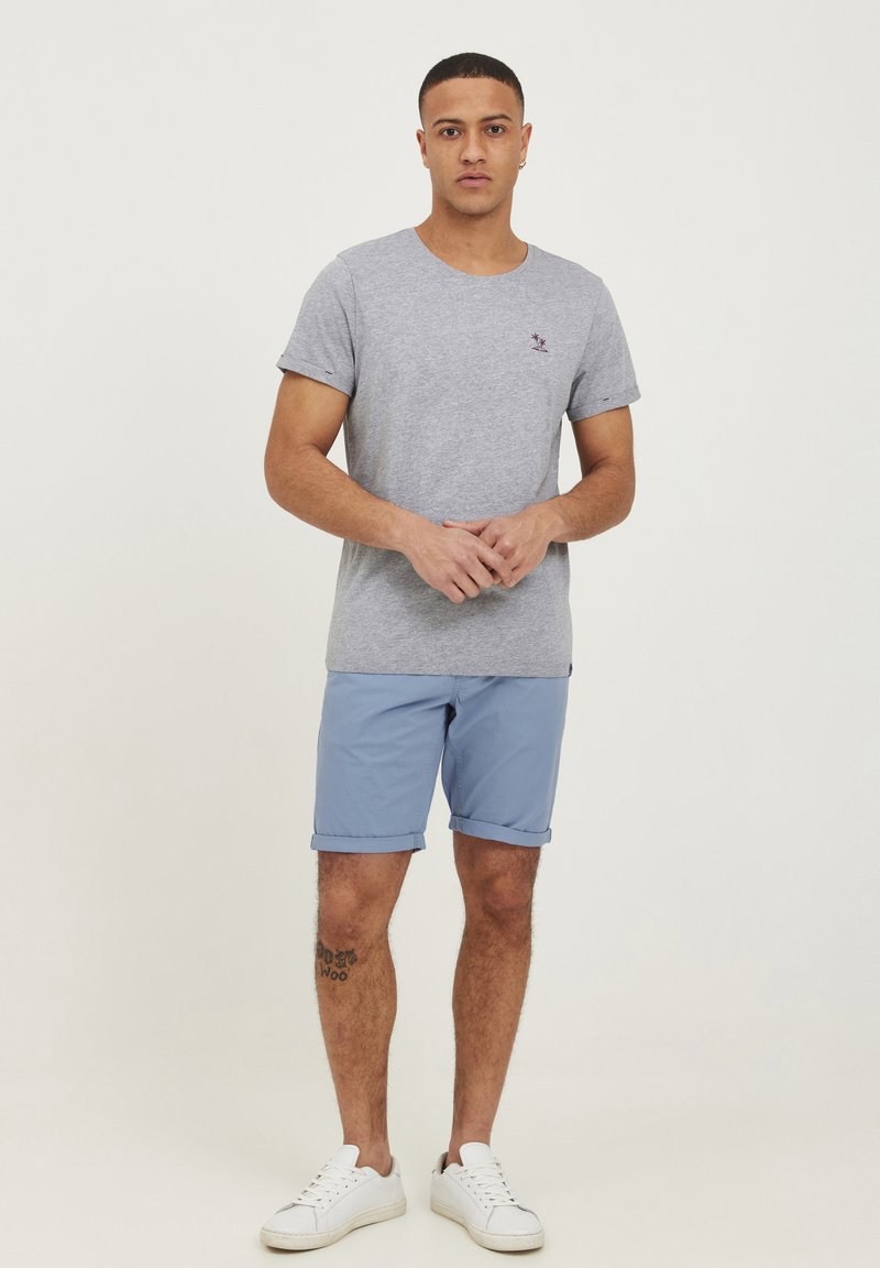 Tricou gri cu mânecă scurtă și guler rotund, având un mic logo brodat; asociat cu pantaloni scurți bleu cu mansete rulate și adidași albi.
