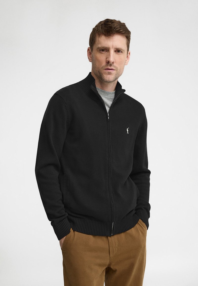 Uomo che indossa un maglione nero con cerniera e piccolo logo ricamato, maglietta grigia sotto e pantaloni marroni, in piedi davanti a uno sfondo semplice.