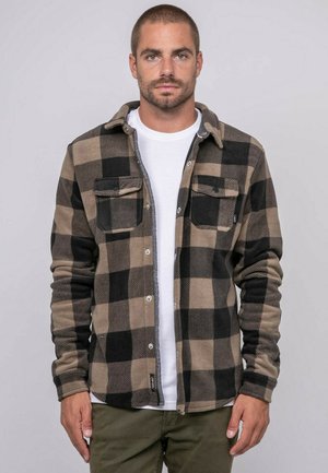 Homme debout, faisant face à l'avant, portant une veste à carreaux beige et noire boutonnée par-dessus une chemise blanche et un pantalon olive, sur un fond uni.