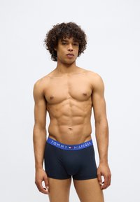 Fit muškarac s kovrčavom kosom nosi mornarsko plave Tommy Hilfiger bokserice s plavim pojasom, stoji naspram obične bijele pozadine.