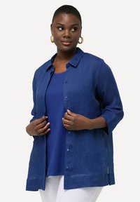 Camisa de lino azul con cuello, mangas tres cuartos, cierre de botones y aberturas laterales, superpuesta sobre una camiseta azul y combinada con pantalones blancos.