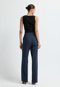 Vrouw met een bun, gekleed in een zwarte mouwloze top en marineblauwe gestreepte broek, staand met de rug naar de camera op een witte achtergrond.