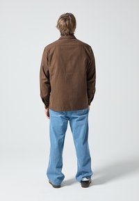 Smeđa košulja s dugmadima i klasičnim ovratnikom, vidljiva tekstura tkanine, u kombinaciji sa svijetloplavim trapericama, prikazana s leđa.