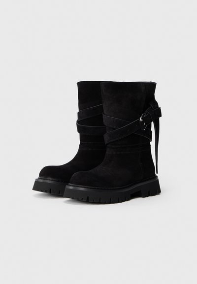 Botas de ante negras hasta la mitad de la pantorrilla con suelas gruesas de llanta y correas envolventes que presentan hebillas de plata sobre un fondo claro y sencillo.