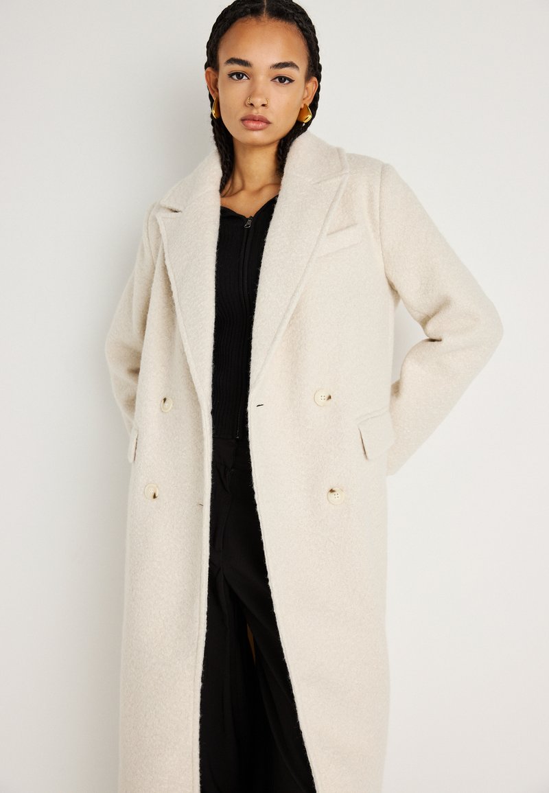 Krémový, dvouřadý kabát z měkkého a texturovaného materiálu. Má klopové límce, postranní kapsy a světlé knoflíky. Hladká vnitřní podšívka.