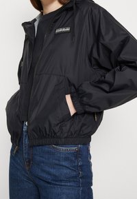 Cazadora negra con capucha, puños elásticos y dobladillo. Cuenta con un zip frontal y un parche con el logo. Combinada con jeans azules.