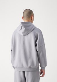 Grå huvtröja med avslappnad passform, ribbade ärmslut och nederkant. Mjuk, texturerad tyg. Bakifrån visas en enkel, minimalistisk design.